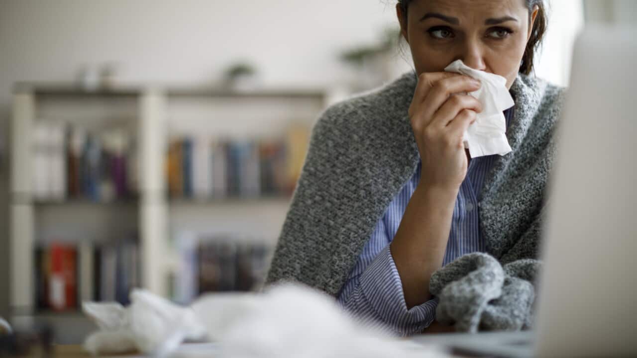 A woman is blowing her nose into a tissue she is holding as she sits in front of a laptop. Books are stacked on a shelf behind her.