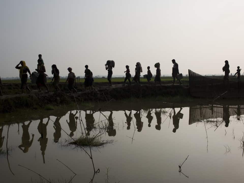 A dozen people walk across path near water, with reflections seen on the water.