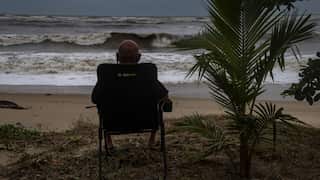 A person sits in a folding chair on a sandy beach, viewed from behind, facing rough ocean waves under a cloudy sky.