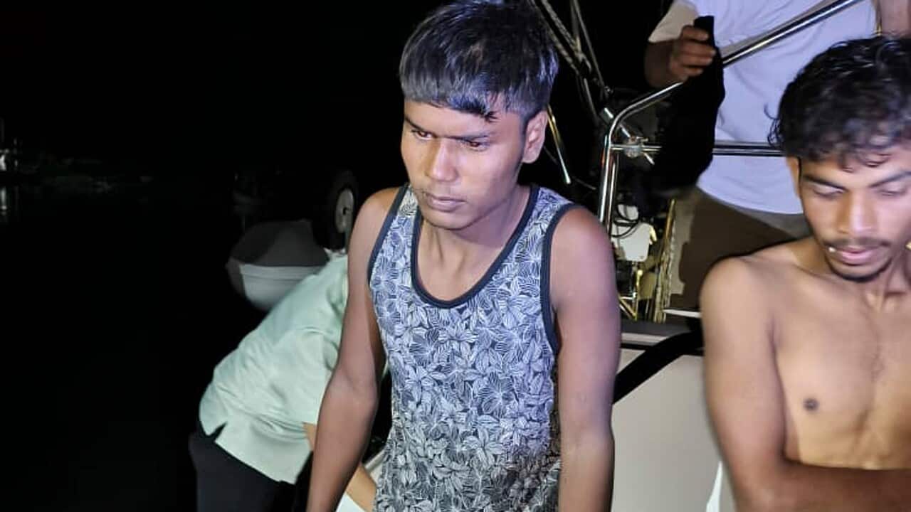 Two men sit in the dark on the edge of a boat, one without a shirt.