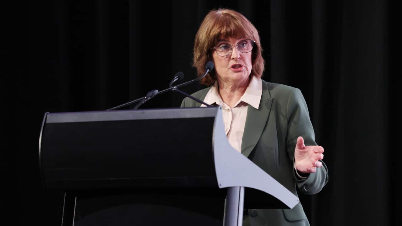 A woman in a green suit jacket and glasses gives a speech at a plinth with a black stage behind her