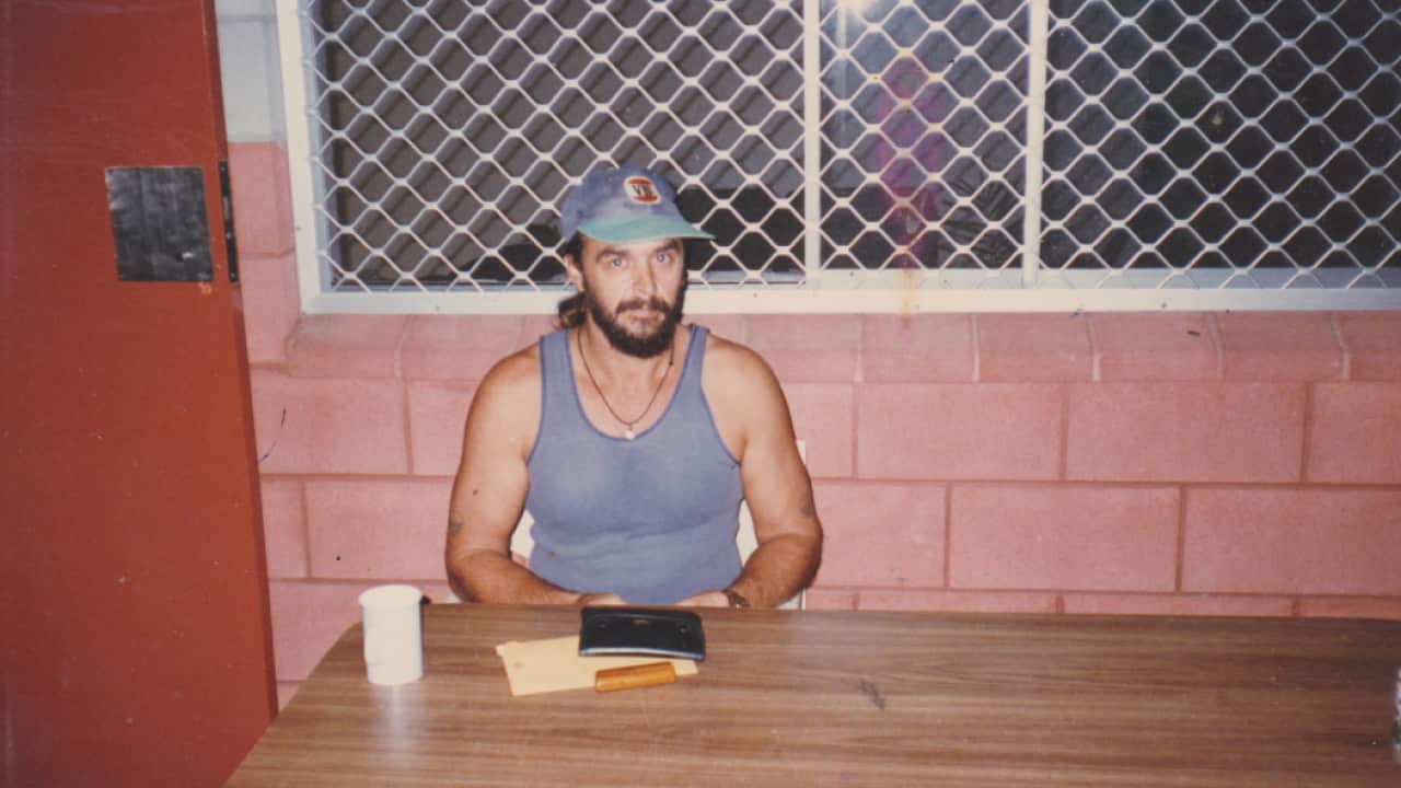 A bearded man wearing a blue vest and basketball cap sits at a table in a prison.