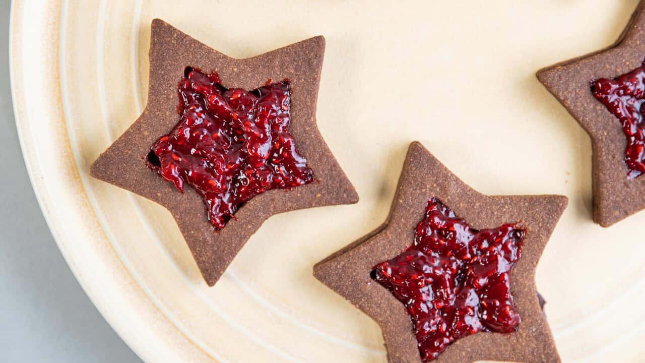 Seen from overhead, two star-shaped chocolate biscuis sit on a round cream-coloured plate. Each star is filled with vibrant strawberry jam. The edge of two more biscuits can also be seen.