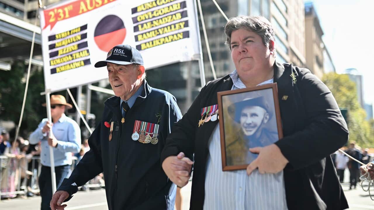 Two people - one elderly, one younger - both wearing blazers with Army medals, holding hands as they march down a street. One is carrying a photo frame showing a young soldier.