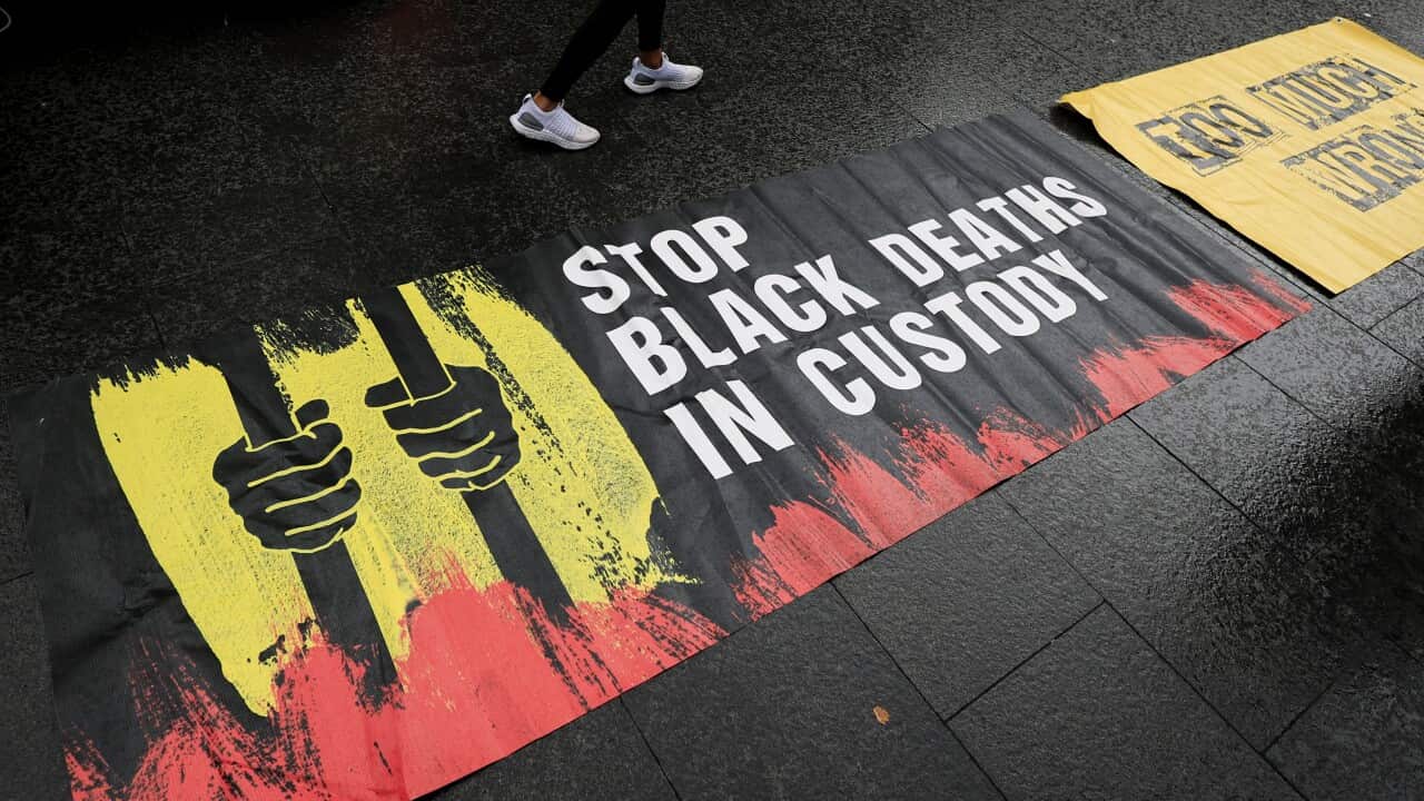 A photo of a protest banner, laid out on pavement. The banner depicts two First Nations hands gripping prison bars in the red, yellow and black colours of the Aboriginal flag. The banner reads "Stop black deaths in custody."