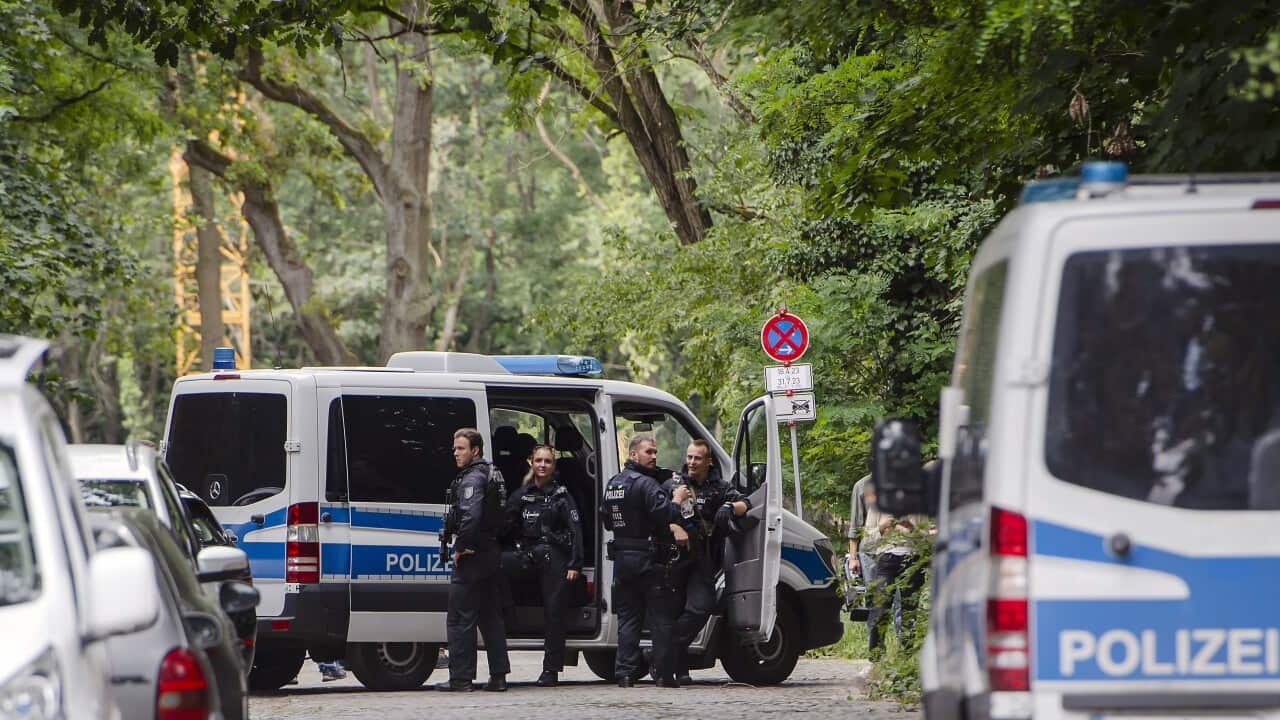Blue and white police vans parked on the side of a gravel road, surrounded by leafy trees. Four policemen in uniform gather outside the vans.