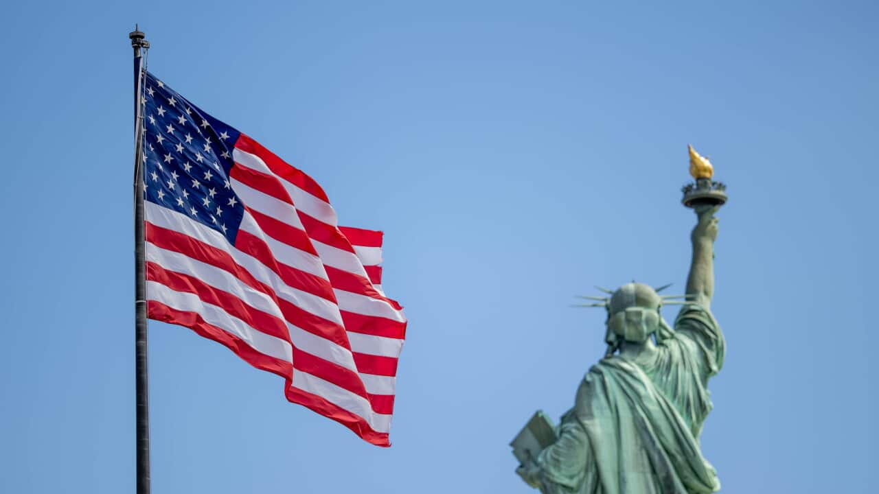 An American flag flutters in the foreground on the left, while the Statue of Liberty is seen from behind and softly out of focus against a clear blue sky on the right.