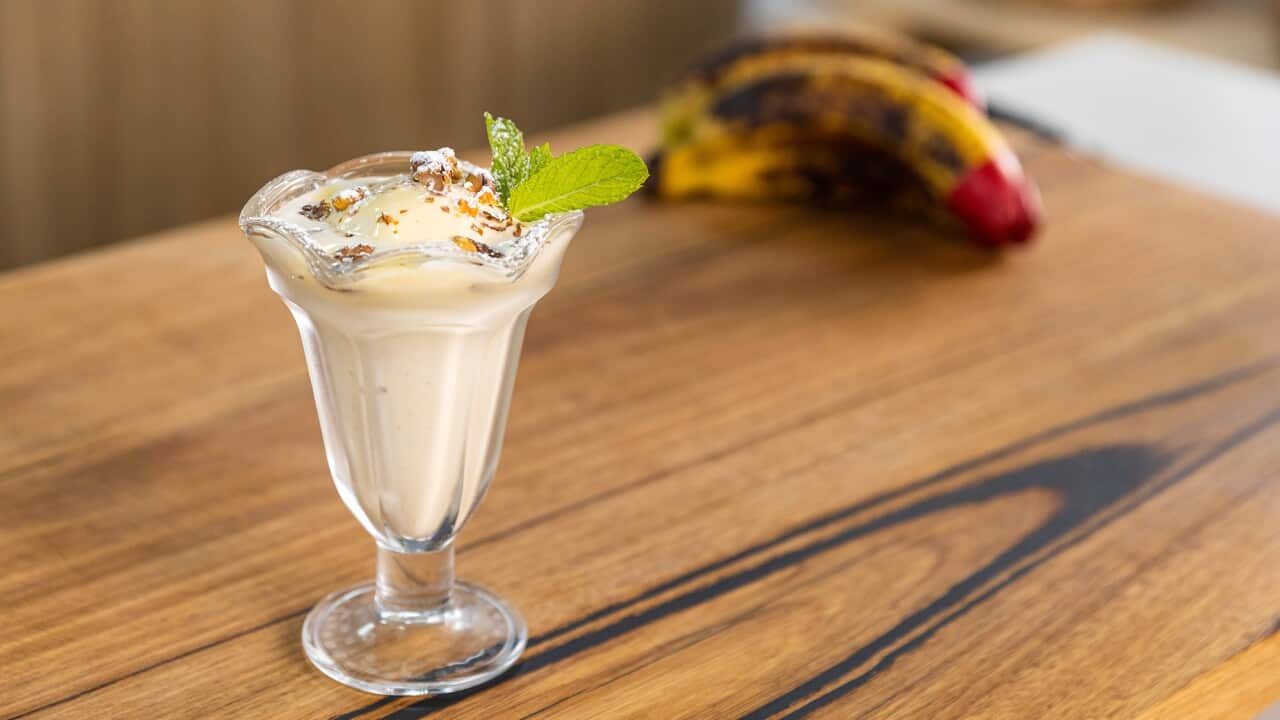 A sundae-style glass sits on a wooden surface. It is filled with a pale, creamy gelato and topped with nuts.
