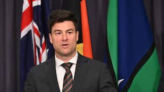 A man in a dark suit, white shirt, and striped tie speaks at a press conference podium, with the Australian flag and several Aboriginal and Torres Strait Islander flags displayed behind him against a dark curtain.