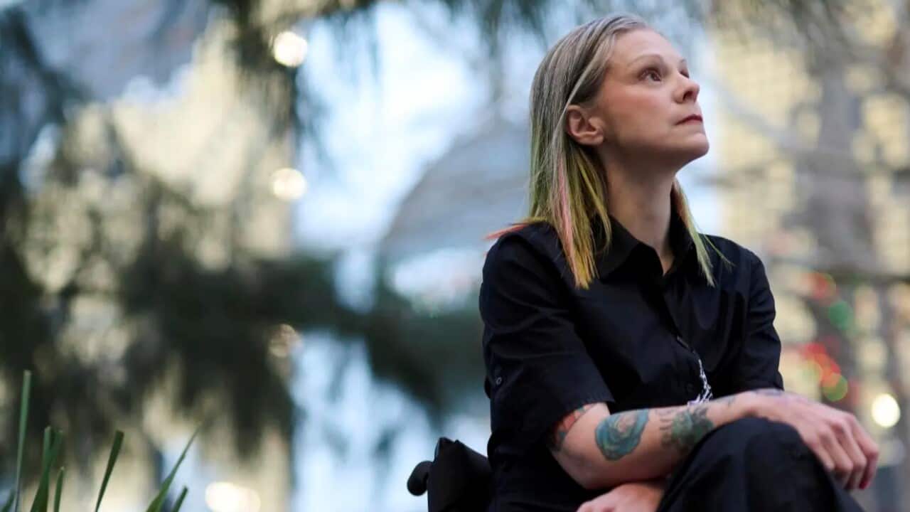 A woman in a wheelchair dressed in dark clothing with colourful hair looking off into the distance