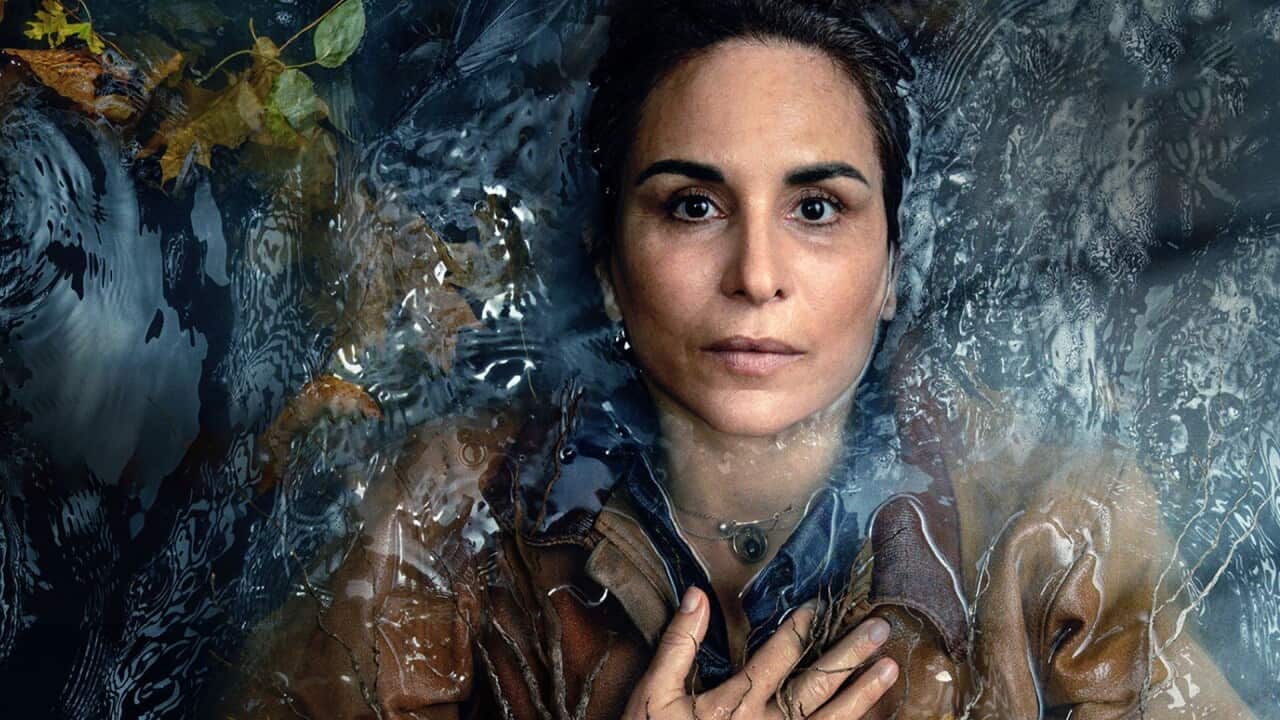 The head and shoulders of a woman lying in rippling water are seen from overhead; she stares straight up with a slightly haunted expression, and one hand on her chest.