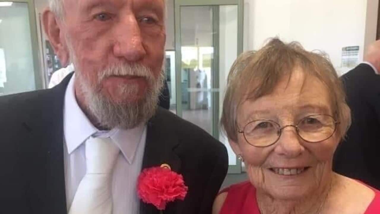 An elderly man in a black suit with a pink flower on his lapel next to an elderly woman in a pink dress.