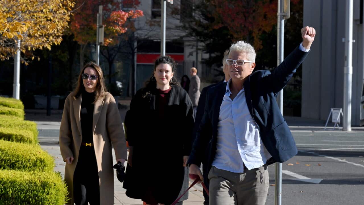 A man wearing a blue jacket and white glasses raises his fist as he walks with two women walking behind him.