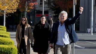 A man wearing a blue jacket and white glasses raises his fist as he walks with two women walking behind him.