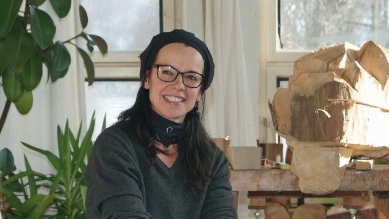 The artist Ilona Herreiner sits on a block of wood in an art studio.