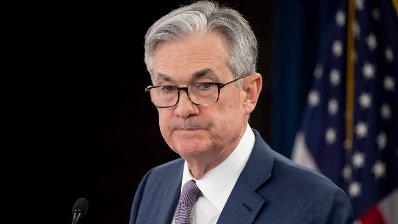 A man with grey hair, wearing a blue suit, stands in front of a microphone with his lips pursed.