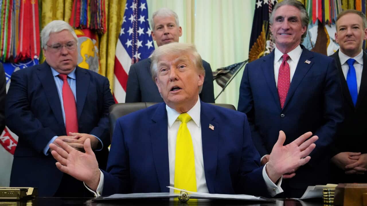 Donald Trump in a blue suit and yellow tie sits at his desk in the Oval Office surrounded by old, white, smiling men.