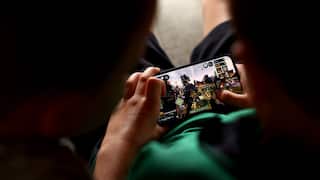 Two children play Roblox on an iPhone. The image is taken over their shoulders, the screen placed between them. One child watches the other play.
