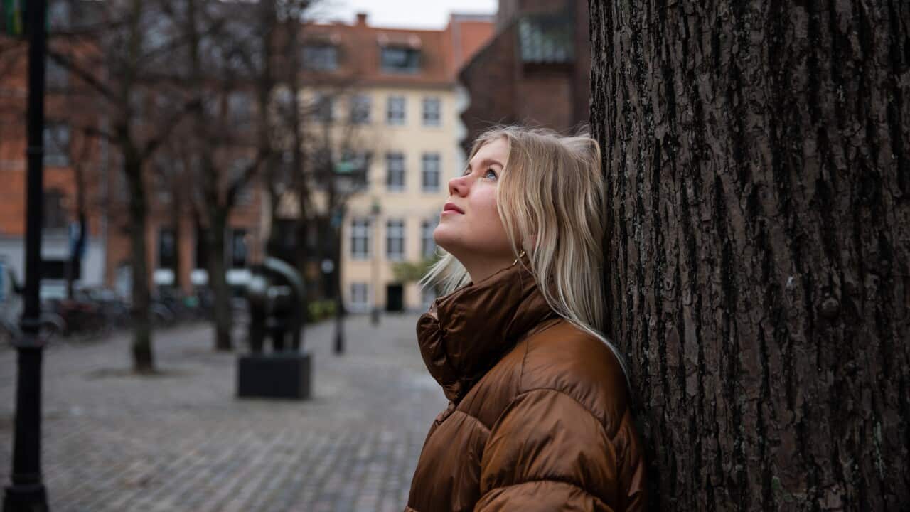 A woman with blonde hair leans up against a tree.