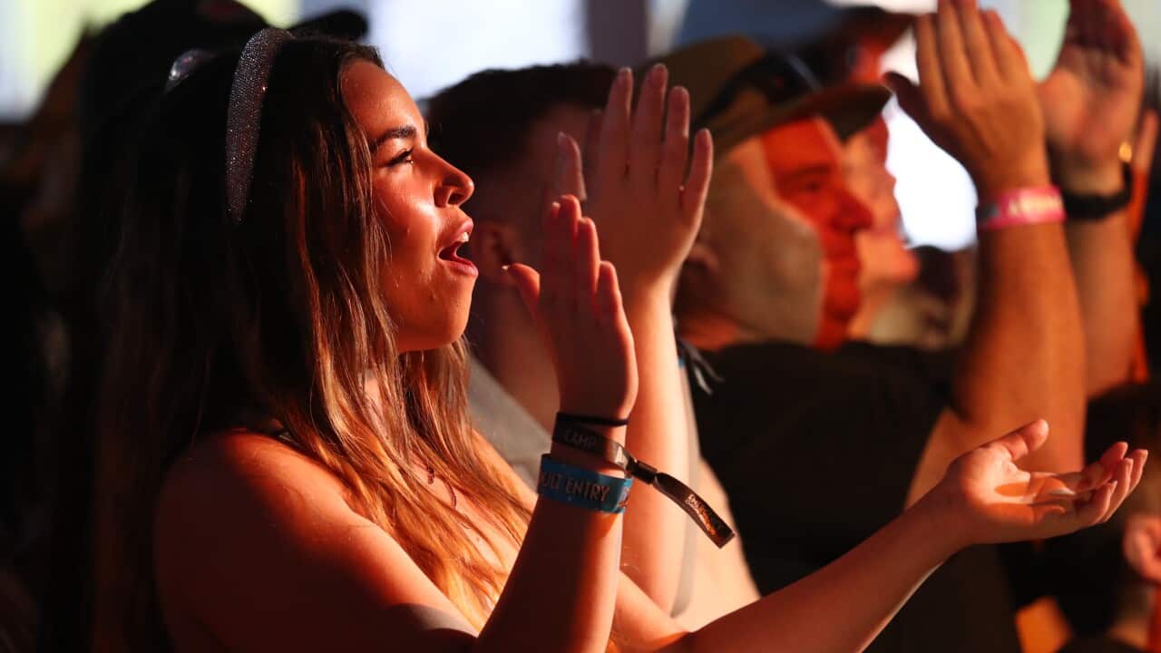 A row of smiling, happy people in an audience stare up towards a stage, holding their hands up in applause.
