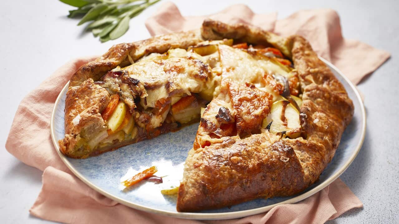 A round, rustic pie with folded over pastry sits on a blue plate, on a peach-toned napkin. A slice has been removed, showing the colourful filling. There is golden cooked cheese on the top of the filling, where it is not covered by the folded-in pastry crust.