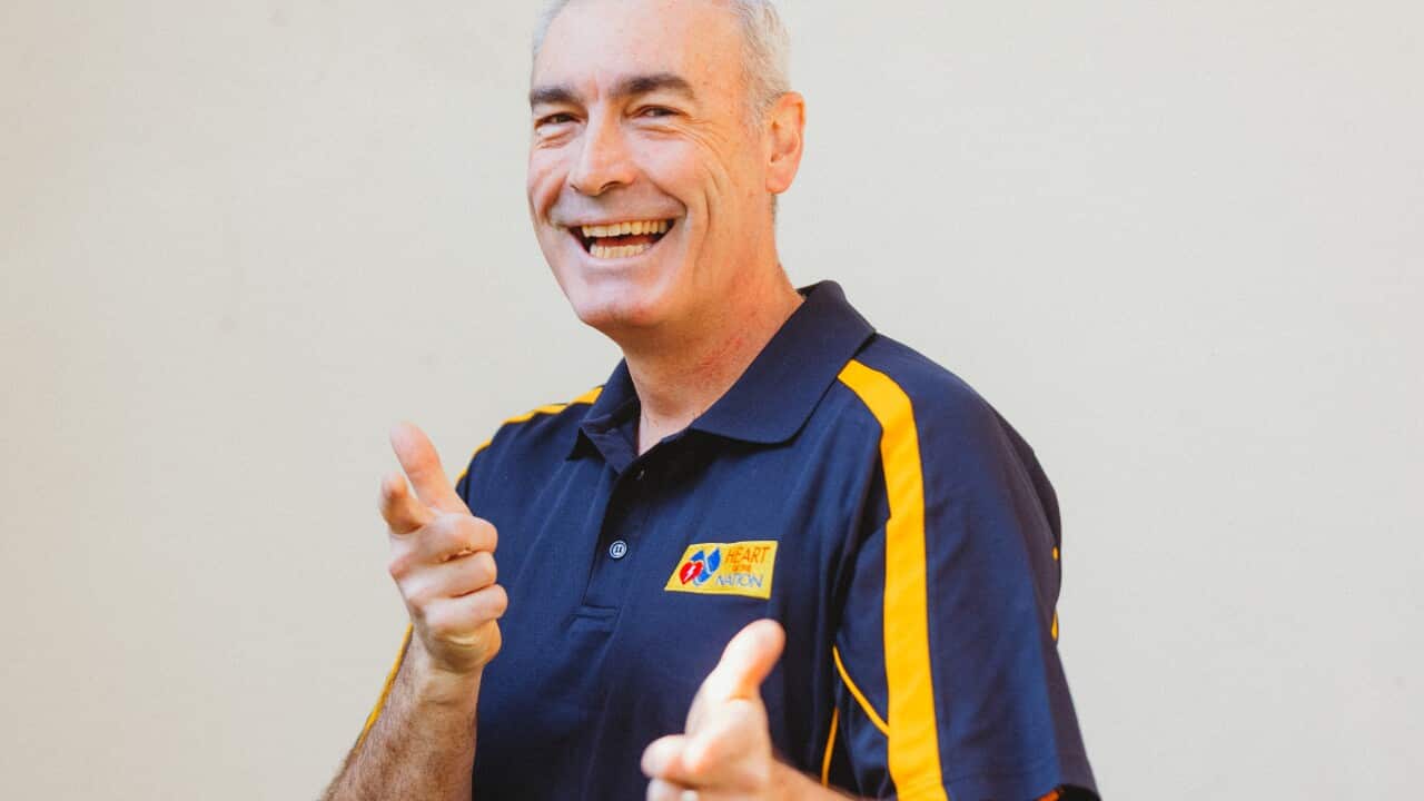 A man wearing a dark blue polo shirt with yellow stripes smiles with both of his index fingers pointed towards the camera.