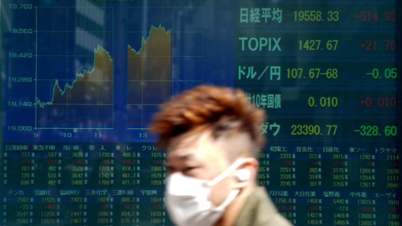 A man wearing a protective mask walks past an electronic stock board showing Japan's Nikkei 225 index.
