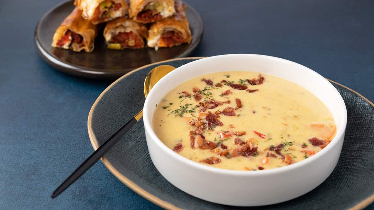 A bowl of yellow soup sits on a blue plate; behind, rolls sit on another plate.