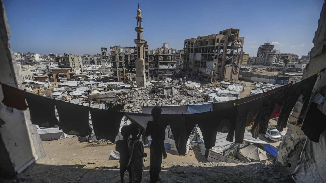 View from a damaged building over a densely packed tent camp and widespread urban destruction, with ruined buildings, a damaged mosque minaret and three children silhouetted in the foreground.