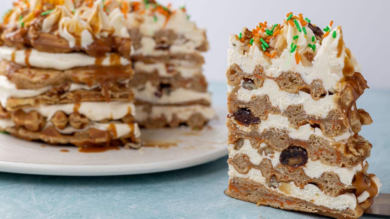 A cake made with five layers of cake and icing sits on a white platter. A piece as been cut from the cake and sits at the front, to one side. The top of the cake is decorated with piped peaks of icing and coloured sprinkles. Caramel sauce can be seen on the sides of the cake.