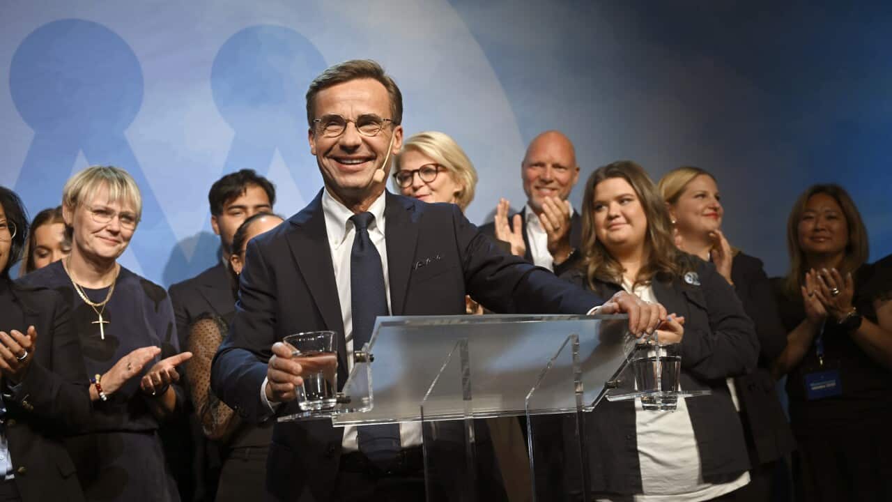 A man stands at a lectern surrounded by people smiling and clapping