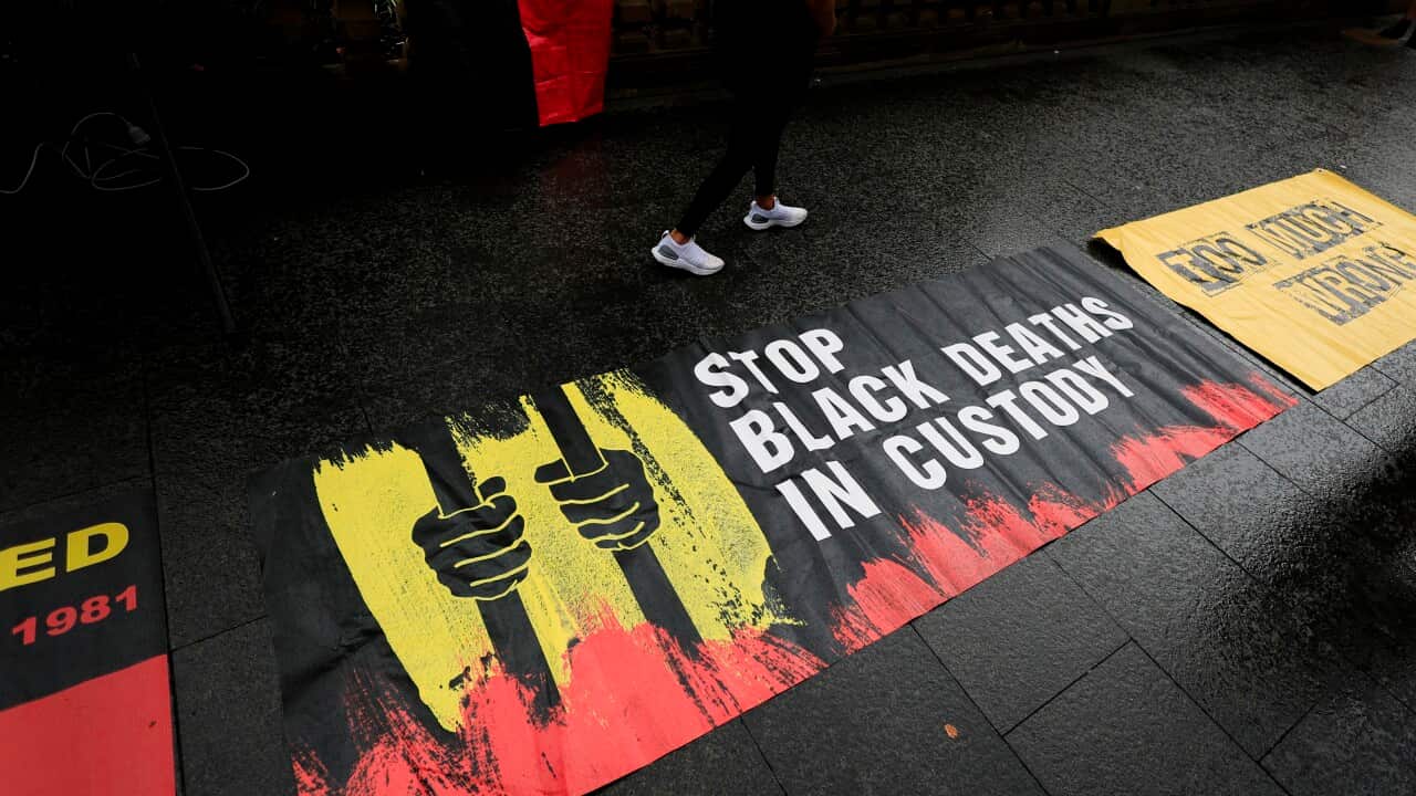 A photo of a protest banner, laid out on pavement. The banner depicts two First Nations hands gripping prison bars in the red, yellow and black colours of the Aboriginal flag. The banner reads “Stop black deaths in custody.“
