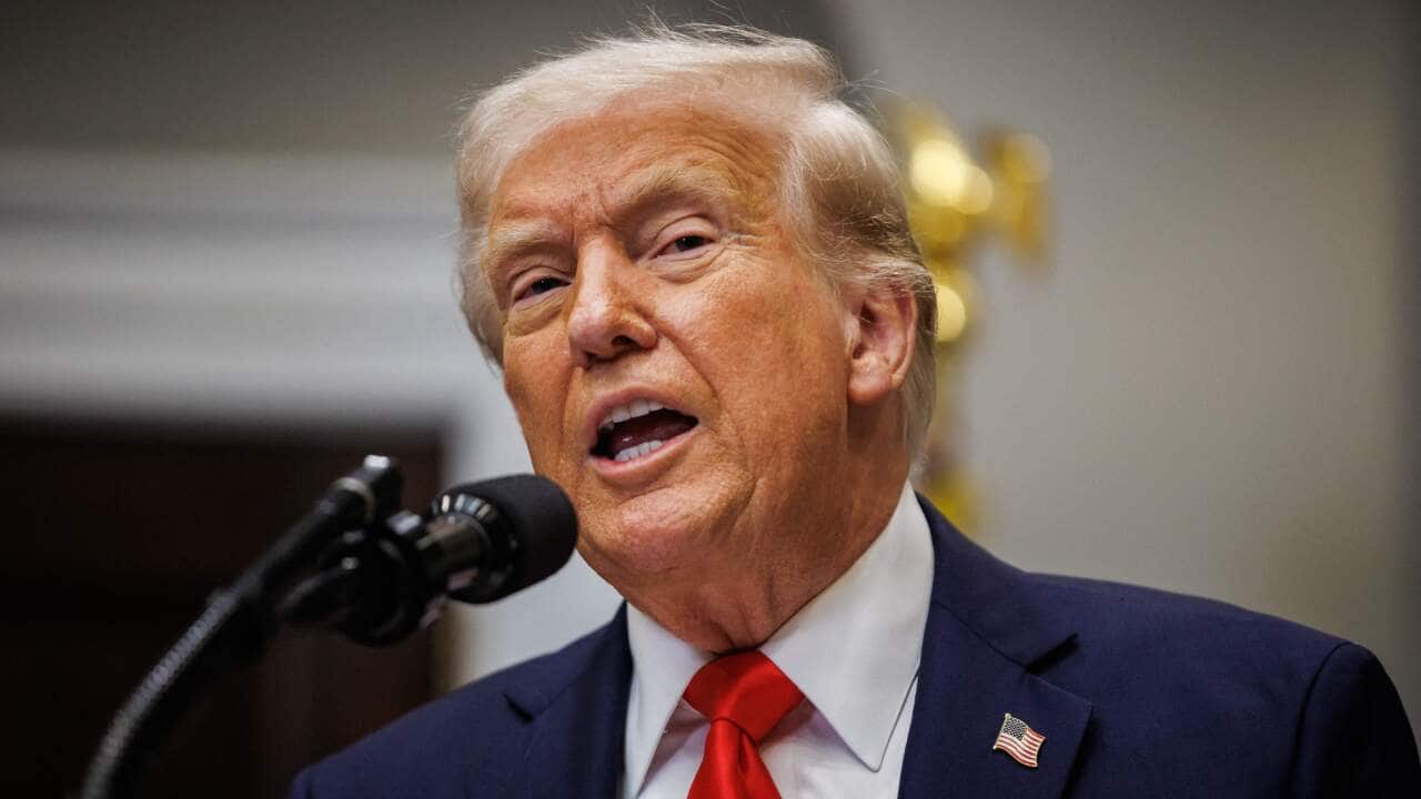 Donald Trump, in a blue suit and red tie, is speaking into a microphone.