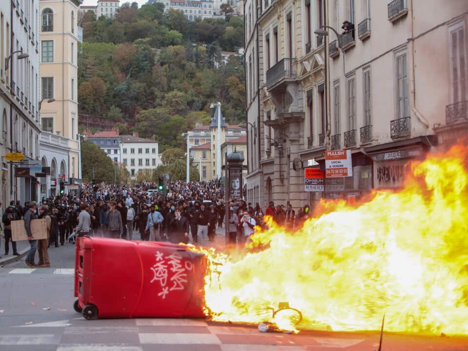 A large street protest fills a city road lined with multi-storey historic buildings. In the foreground, an overturned red rubbish bin lies on its side near a road intersection while a large fire burns across part of the street, producing bright yellow and orange flames. A dense crowd of hundreds of people stretches into the distance behind the fire, occupying the width of the roadway. Shopfronts, streetlights and apartment balconies line both sides of the street, and a tree-covered hillside rises in the background behind the urban streetscape. Visible signs on buildings include “MALSH”, “BREQUILLE” and a real estate advertisement.