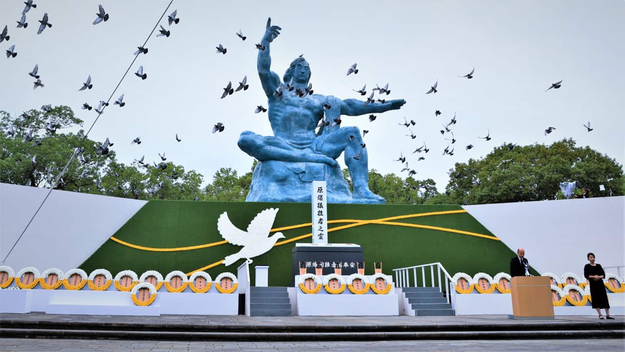 Doves fly over the large blue Peace Statue during a memorial ceremony in Nagasaki, Japan, featuring floral memorial wreaths along a stage and a speaker at a podium.