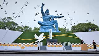 Doves fly over the large blue Peace Statue during a memorial ceremony in Nagasaki, Japan, featuring floral memorial wreaths along a stage and a speaker at a podium.