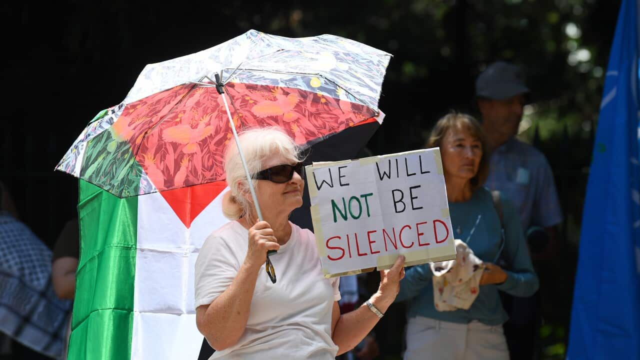 A woman holding an umbrella and a sign that says “We will not be silenced“. Other people are standing around here. A Palestinian flag is also visible behind her.