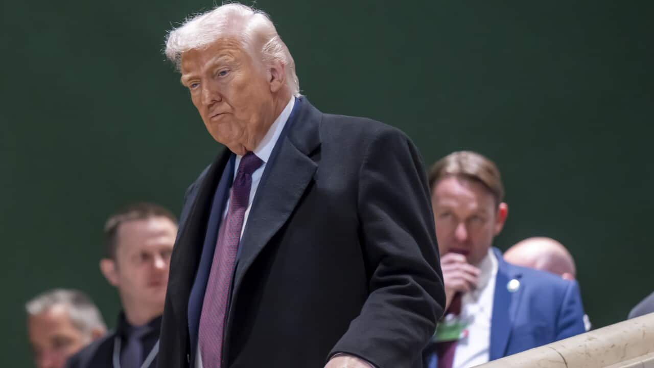 A man in a dark suit with a red tie and white hair walks down a set of stairs