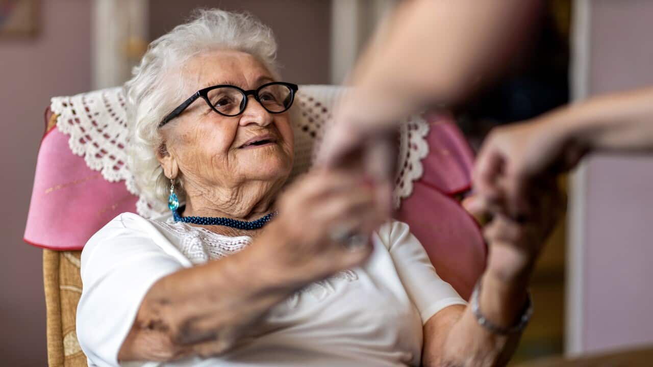 Female home carer supporting old woman to stand up from the armchair at care home