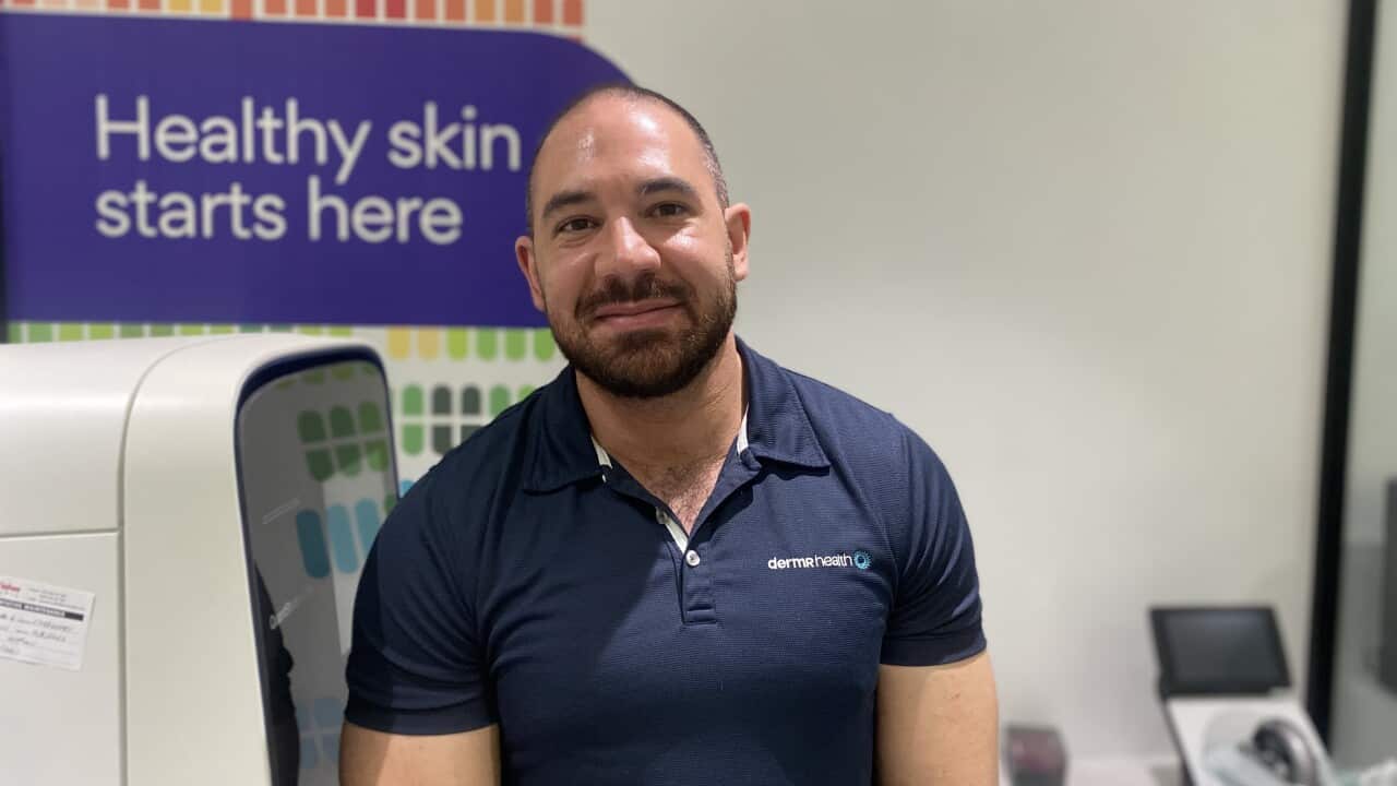 A man in a blue t-shirt is smiling in front of a 'Healthy skin starts here' poster.
