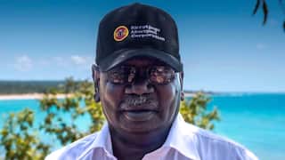 An Indigenous man wearing a black ht nd sunglasses in front of a blue ocean