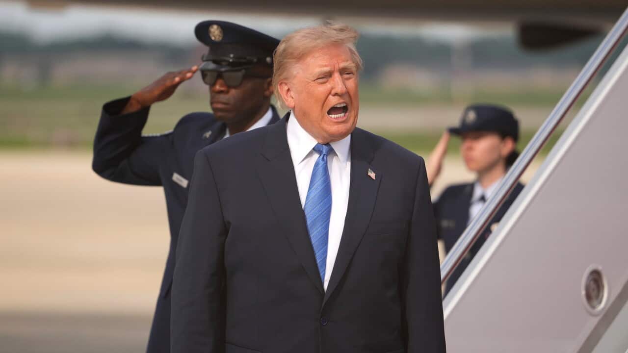 Donald Trump, wearing a suit, speaking next to a set of stairs, with two soldiers behind him saluting him.