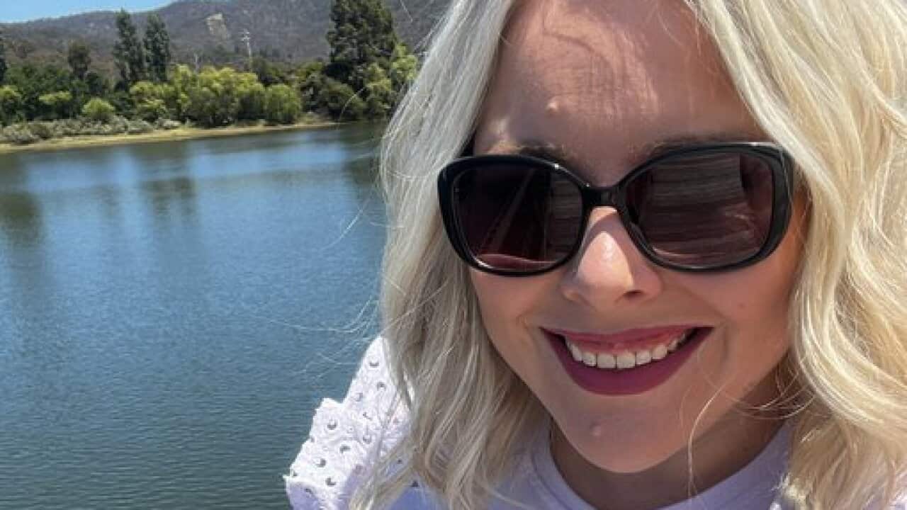 A smiling woman, wearing sunglasses in front of a lake and hills.