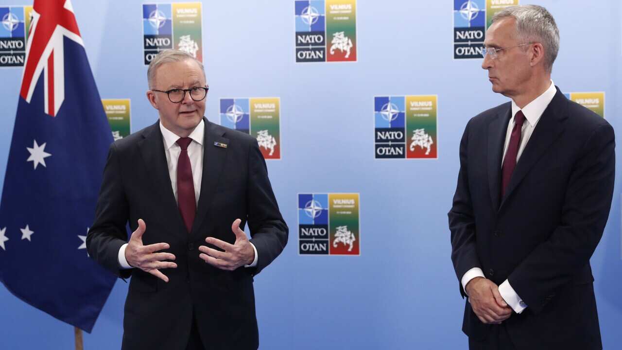 Two men in black suits stand in front of a blue wall.
