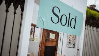 A sign for a recently sold house sits against a beige picket fence. The sign bears a picture of a red brick and wood fireplace.