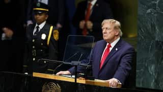 Donald Trump, in a dark suit and red tie stands at a United Nations lectern, speaking behind a transparent shield, with uniformed security personnel visible in the background. The UN emblem is displayed on the front of the podium.