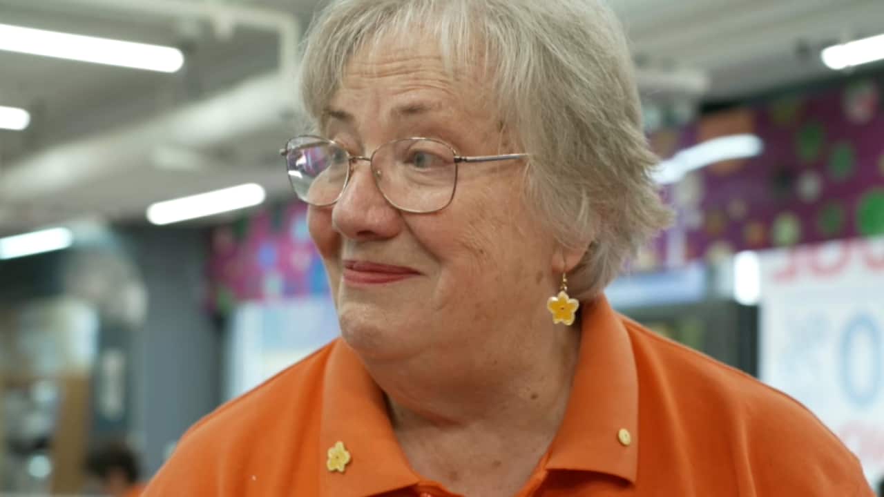 An elderly woman with short grey hair and glasses smiles warmly while wearing an orange polo shirt adorned with yellow flower pins.
