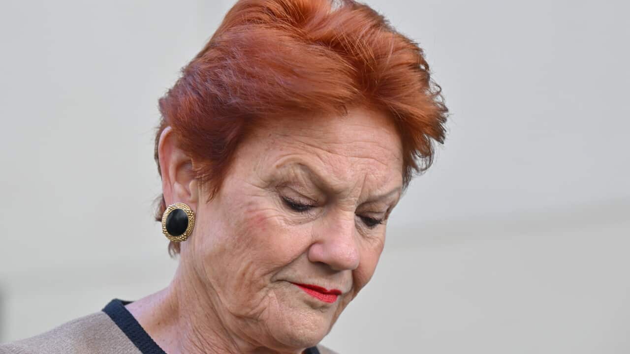 Close-up portrait of an older woman with short red hair, red lipstick and large earrings, looking downward against a blurred light-coloured background.