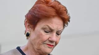 Close-up portrait of an older woman with short red hair, red lipstick and large earrings, looking downward against a blurred light-coloured background.