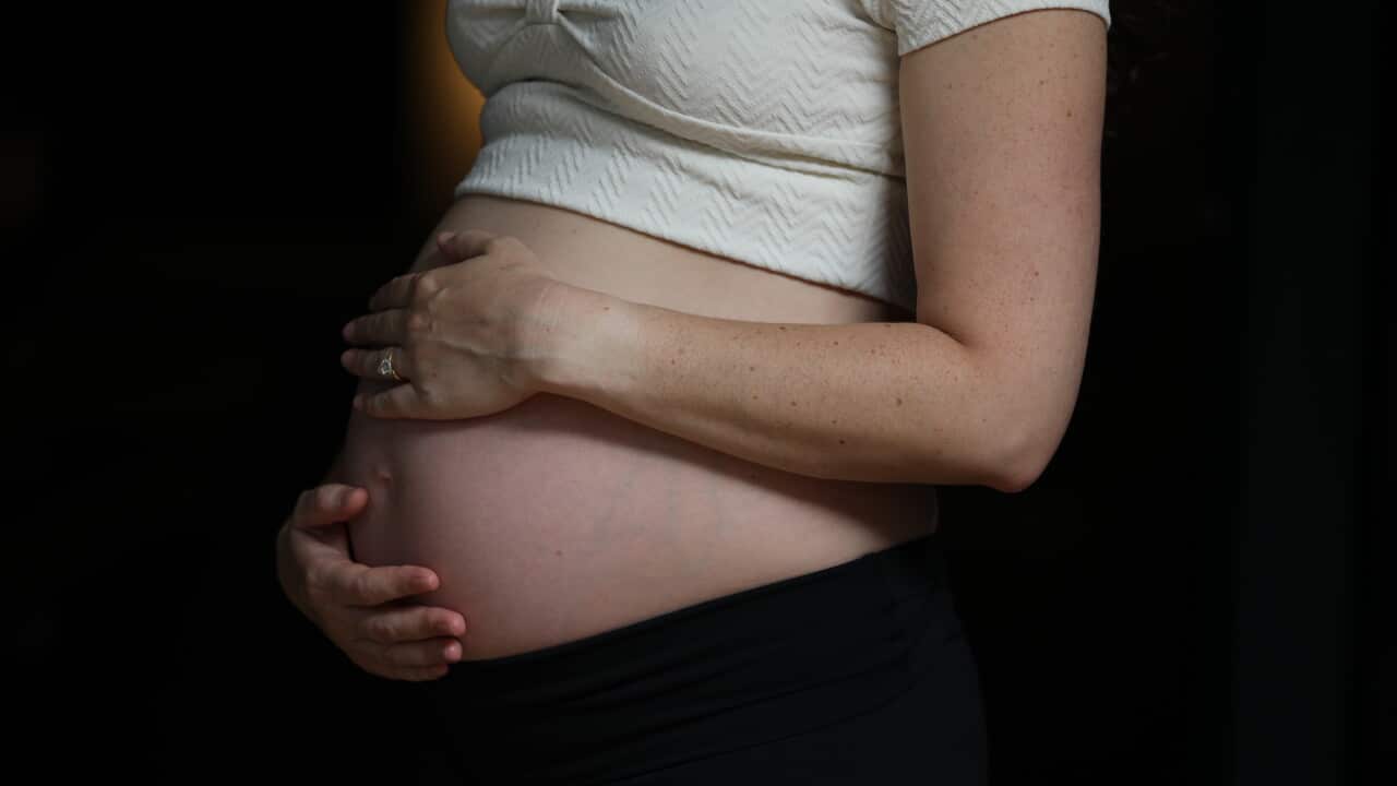 A pregnant woman holds her belly wearing a white shirt and black pants against a dark background